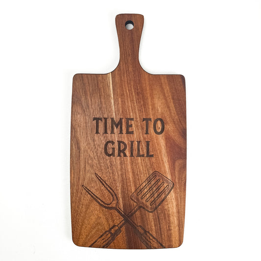 Wooden cutting board with 'Time to Grill' engraving and grill tools illustration on a white background. Acacia wood laser engraved charcuterie board and serving board handmade in Spain 