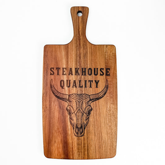 Wooden cutting board with a bull skull design and 'Steakhouse Quality' text on a white background. Acacia wood laser engraved charcuterie board and serving board handmade in Spain 