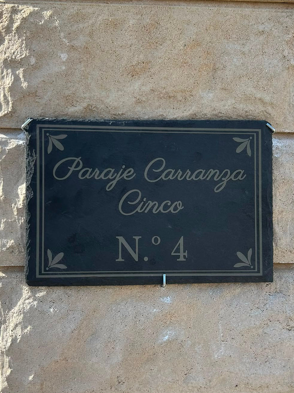 A dark slate rectangular house sign with white script reading 'Paraje Carranza Cinco N.° 4' mounted on a light beige stone pillar outdoors.