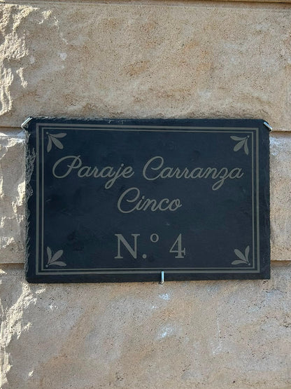 A dark slate rectangular house sign with white script reading 'Paraje Carranza Cinco N.° 4' mounted on a light beige stone pillar outdoors.